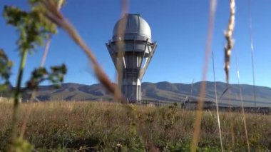 Large buildings of the observatory in the form of a dome. The telescopes are closed. Beautiful tall yellow-green grass develops in the wind. Blue sky and green hills in the distance. The Assy Plateau