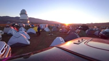 The time lapse of dawn in the camp near the observatory. The sun comes out from behind high hills. Red-yellow color. Lots of tents and cars. Guy launches the drone. Telescopes are covered with domes