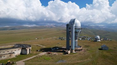 The Assy-Turgen Observatory is high in the mountains. There is a tent camp next to the observatory. Large cumulus clouds in a blue sky. Yellow-green hills, forest in places. Top view from a drone