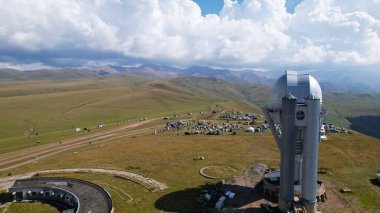 The Assy-Turgen Observatory is high in the mountains. There is a tent camp next to the observatory. Large cumulus clouds in a blue sky. Yellow-green hills, forest in places. Top view from a drone
