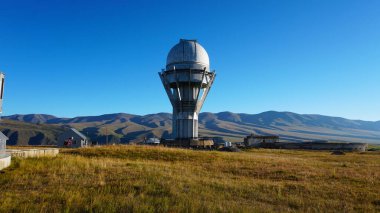 A large observatory is protected by a barbed fence. There is a long sharp wire around perimeter. Buildings with domes and a telescope are visible in distance. Yellow-green grass. Mountainous terrain
