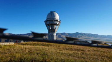 A large observatory is protected by a barbed fence. There is a long sharp wire around perimeter. Buildings with domes and a telescope are visible in distance. Yellow-green grass. Mountainous terrain
