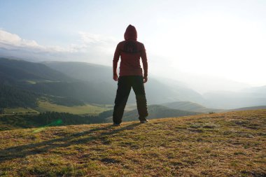 A guy on the edge of the hill admires the view. In the distance you can see high mountains and green hills covered with forest. Wide margins. Clouds are visible and sun is shining brightly. Kazakhstan