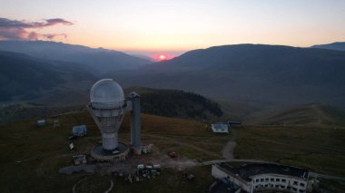 Two large telescope domes at sunset. Drone view of Assy-Turgen Observatory. Beautiful red sunset. Green hills and clouds. Tourists watch the sun. There is a large tent camp and cars nearby. Kazakhstan
