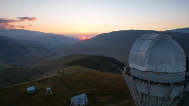 Two large telescope domes at sunset. Drone view of Assy-Turgen Observatory. Beautiful red sunset. Green hills and clouds. Tourists watch the sun. There is a large tent camp and cars nearby. Kazakhstan