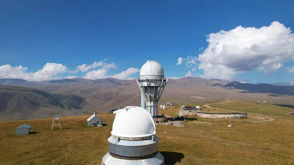 The Assy-Turgen Observatory is high in the mountains. There is a tent camp next to the observatory. Large cumulus clouds in a blue sky. Yellow-green hills, forest in places. Top view from a drone