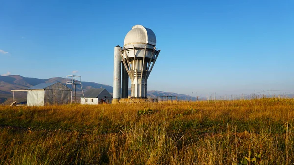 Large buildings of the observatory in the form of a dome. The telescopes are closed. Beautiful tall yellow-green grass develops in the wind. Blue sky and green hills in the distance. The Assy Plateau