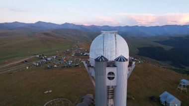 Two large telescope domes at sunset. Drone view of Assy-Turgen Observatory. Beautiful red sunset. Green hills and clouds. Tourists watch the sun. There is a large tent camp and cars nearby. Kazakhstan