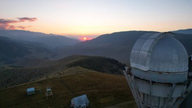 Two large telescope domes at sunset. Drone view of Assy-Turgen Observatory. Beautiful red sunset. Green hills and clouds. Tourists watch the sun. There is a large tent camp and cars nearby. Kazakhstan