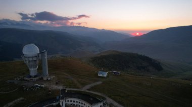 Two large telescope domes at sunset. Drone view of Assy-Turgen Observatory. Beautiful red sunset. Green hills and clouds. Tourists watch the sun. There is a large tent camp and cars nearby. Kazakhstan