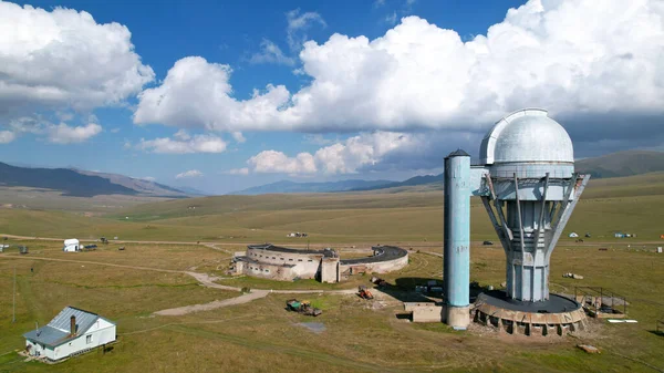 The Assy-Turgen Observatory is high in the mountains. There is a tent camp next to the observatory. Large cumulus clouds in a blue sky. Yellow-green hills, forest in places. Top view from a drone