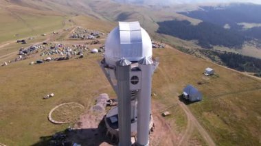 The Assy-Turgen Observatory with a large telescope. A tent camp is located nearby. There are colored tents, cars and people are resting. Big clouds over green hills. Two domes for studying the stars