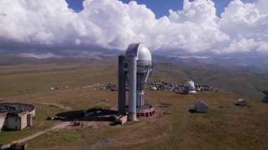 The Assy-Turgen Observatory with a large telescope. A tent camp is located nearby. There are colored tents, cars and people are resting. Big clouds over green hills. Two domes for studying the stars
