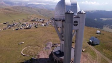 The Assy-Turgen Observatory with a large telescope. A tent camp is located nearby. There are colored tents, cars and people are resting. Big clouds over green hills. Two domes for studying the stars