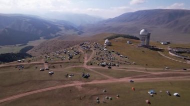 The Assy-Turgen Observatory with a large telescope. A tent camp is located nearby. There are colored tents, cars and people are resting. Big clouds over green hills. Two domes for studying the stars