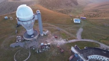 Two large telescope domes at sunset. Drone view of Assy-Turgen Observatory. Beautiful red sunset. Green hills and clouds. Tourists watch the sun. There is a large tent camp and cars nearby. Kazakhstan