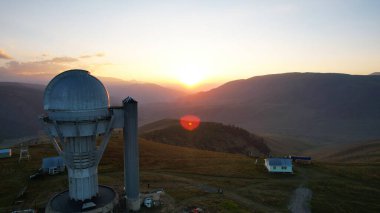 Two large telescope domes at sunset. Drone view of Assy-Turgen Observatory. Beautiful red sunset. Green hills and clouds. Tourists watch the sun. There is a large tent camp and cars nearby. Kazakhstan