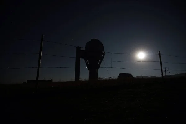 A big bright moon in the starry sky. The observatory is fenced with a barbed fence. The dome above the building with a large telesk. The yellow-green grass is trampled. There are several houses