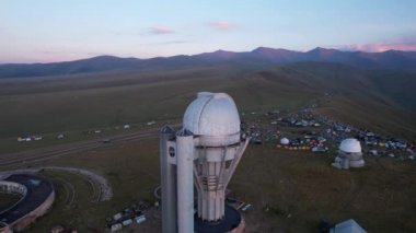 Two large telescope domes at sunset. Drone view of Assy-Turgen Observatory. Beautiful red sunset. Green hills and clouds. Tourists watch the sun. There is a large tent camp and cars nearby. Kazakhstan