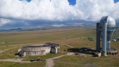 The Assy-Turgen Observatory is high in the mountains. There is a tent camp next to the observatory. Large cumulus clouds in a blue sky. Yellow-green hills, forest in places. Top view from a drone