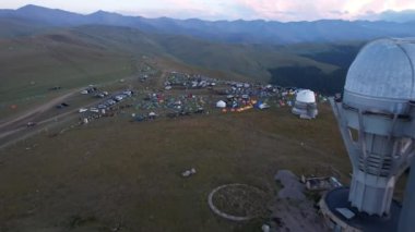 Two large telescope domes at sunset. Drone view of Assy-Turgen Observatory. Beautiful red sunset. Green hills and clouds. Tourists watch the sun. There is a large tent camp and cars nearby. Kazakhstan