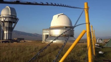 A large observatory is protected by a barbed fence. There is a long sharp wire around perimeter. Buildings with domes and a telescope are visible in distance. Yellow-green grass. Mountainous terrain