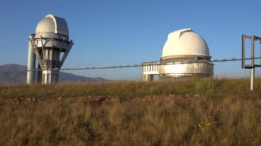 A large observatory is protected by a barbed fence. There is a long sharp wire around perimeter. Buildings with domes and a telescope are visible in distance. Yellow-green grass. Mountainous terrain