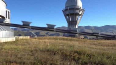 A large observatory is protected by a barbed fence. There is a long sharp wire around perimeter. Buildings with domes and a telescope are visible in distance. Yellow-green grass. Mountainous terrain
