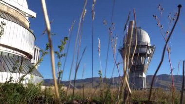 Large buildings of the observatory in the form of a dome. The telescopes are closed. Beautiful tall yellow-green grass develops in the wind. Blue sky and green hills in the distance. The Assy Plateau