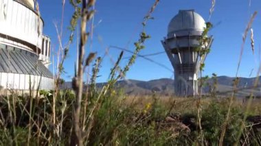 Large buildings of the observatory in the form of a dome. The telescopes are closed. Beautiful tall yellow-green grass develops in the wind. Blue sky and green hills in the distance. The Assy Plateau