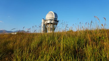 Large buildings of the observatory in the form of a dome. The telescopes are closed. Beautiful tall yellow-green grass develops in the wind. Blue sky and green hills in the distance. The Assy Plateau