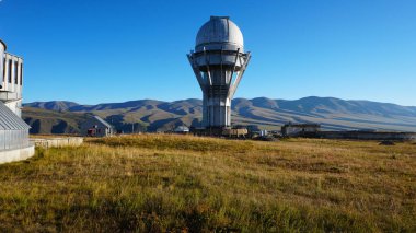 A large observatory is protected by a barbed fence. There is a long sharp wire around perimeter. Buildings with domes and a telescope are visible in distance. Yellow-green grass. Mountainous terrain