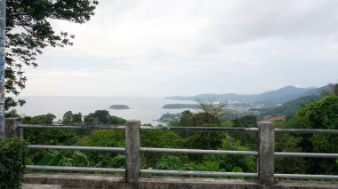 Beautiful view of the island and the sea. Large palm trees grow on the green hills of the island. The sea and clouds are more visible in the distance. There is a fence on the observation deck. Phuket
