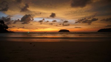 Orange sunset with a view of the sea and the island and the yacht. Green lights are visible in distance, there is a yacht with blue highlights. Clouds are floating. The sea bay. Wet sand reflects sky