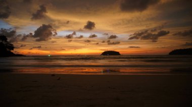 Orange sunset with a view of the sea and the island and the yacht. Green lights are visible in distance, there is a yacht with blue highlights. Clouds are floating. The sea bay. Wet sand reflects sky