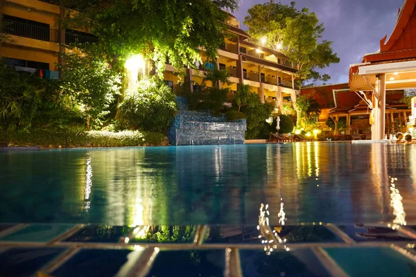 Night pool with lighting and green trees. Smooth water is like a mirror. Lights, trees and buildings are reflected in water. Large balconies. The pool floor is tiled. There are sun loungers. Phuket