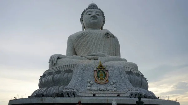 The Big Buddha looks straight into the eyes into the camera. He sits in the lotus position and looks down at people. There is a temple in the center. Gray clouds. Buddha statue in Phuket, Thailand