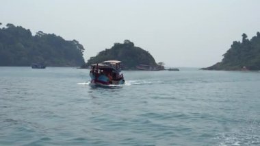 Denizde yüzen eski ahşap Tayland teknesi. Tropikal adalar uzaktan görülebilir, gökyüzü sisli. Turist gemisi denizde dalgalar bırakır. Deniz yoluyla seyahat et. Sarı bir şamandıra yelken açar.