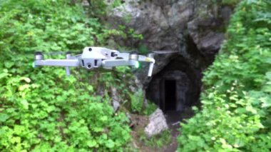 DJI Air2S kuadkopteri mağara girişinin önünde duruyor. Her yerde yeşil çalılar, çimenler ve eğrelti otları var. Dik uçurumlar ve eski bir mağara kapısı. Kazakistan 'ın dağları ve ormanları. Güzel drone