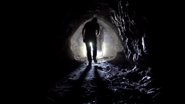 一个拿着手电筒的人从黑暗的洞穴里出来了 灯在隧道的尽头 岩墙和天花板高山上的人造隧道 老旧生锈的门男人的影子 — 图库视频影像