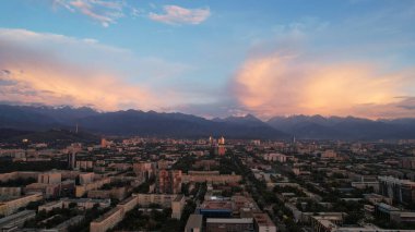 Almaty şehrinin üzerinde bulutlarla destansı bir gün batımı. Bulutların eğimi koyu maviden mor-turuncuya. Yeşil uzun ağaçlar, evler, arabalı yollar. Işıklar açık. Uzakta yüksek dağlar