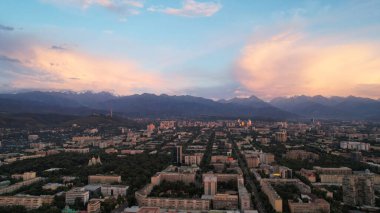 Almaty şehrinin üzerinde bulutlarla destansı bir gün batımı. Bulutların eğimi koyu maviden mor-turuncuya. Yeşil uzun ağaçlar, evler, arabalı yollar. Işıklar açık. Uzakta yüksek dağlar