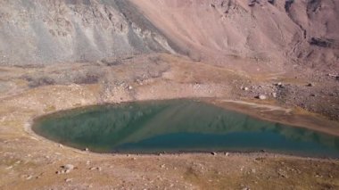Bir dağ gölünün sularının zümrüt rengi. Dağların yansıması olan yeşil bir dağ gölünün drone görüntüsü. Pürüzsüz şeffaf su. Yakınlarda büyük taşlar var. Kuru çimler Kazakistan