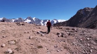 Adam karlı tepeleri gören bir manzarayla dağa tırmanır. Yüksek dağlar mavi gökyüzüne uzanır. Dağların tepeleri buzullarla kaplı. Dik uçurumlar. Büyük taşlar var..