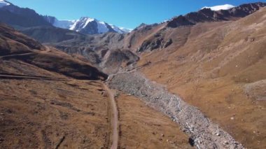 Dağ nehri, karlı tepe manzaralı. Nehir boyunca toprak bir yol var. Dağların yamaçlarında bir sürü taş var. Dağlardaki eski buzullar. Sarı çimenler. Kazakistan