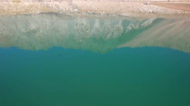 Bir dağ gölünün sularının zümrüt rengi. Dağların yansıması olan yeşil bir dağ gölünün drone görüntüsü. Pürüzsüz şeffaf su. Yakınlarda büyük taşlar var. Kuru çimler Kazakistan