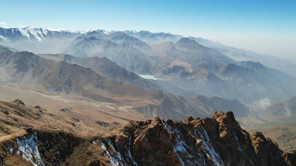 Snowy rocky mountain peaks in a light haze. Aerial view from the drone of the blue sky, light haze and steep cliffs with peaks. A moraine lake can be seen in the distance. Ancient glaciers. Kazakhstan