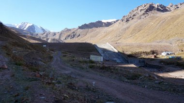 Kayaların arasındaki dağ barajına giden yol. Toprak bir yol evlere ve dağlarda bir çardağa çıkar. Buzullarla kaplı karla kaplı dağlar uzaktan görülebilir. Mavi gökyüzü. Kazakistan