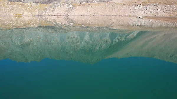 Bir dağ gölünün sularının zümrüt rengi. Dağların yansıması olan yeşil bir dağ gölünün drone görüntüsü. Pürüzsüz şeffaf su. Yakınlarda büyük taşlar var. Kuru çimler Kazakistan