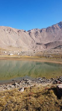 Etrafı kayalarla çevrili şeffaf dağ gölü. Su, kayaları ve eğimi yansıtan bir ayna gibidir. Suyun yüzeyinde hafif dalgalanmalar var. Dibi görünüyor. Büyük taşlar var.